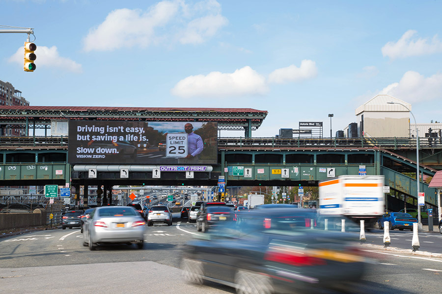 Digital billboard over Astoria Blvd reads, Driving isn’t easy, but saving a life is. Slow down. Features speed limit 25 and traffic below.