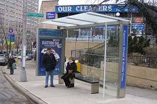 Two people wait under a bus shelter, one is standing and the other is sitting on a bench.