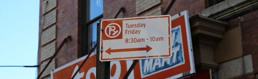 A white and red sign with a P and broom over it indicate street sweeping days.