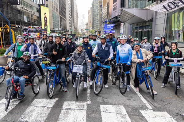 A large group of people wearing bike helmets stand or sit on bicycles and handcycles, ready to ride along a car-free roadway in Manhattan