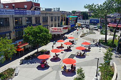 A large public plaza in Queens filled with pedestrians sitting at tables under red umbrellas on a sunny day.