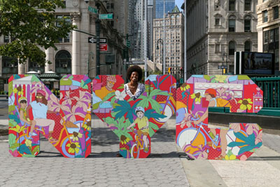 A women stands behind a large sign with the letters N-Y-C covered in colorful designs of people biking, greenery on top of a red and orange background.