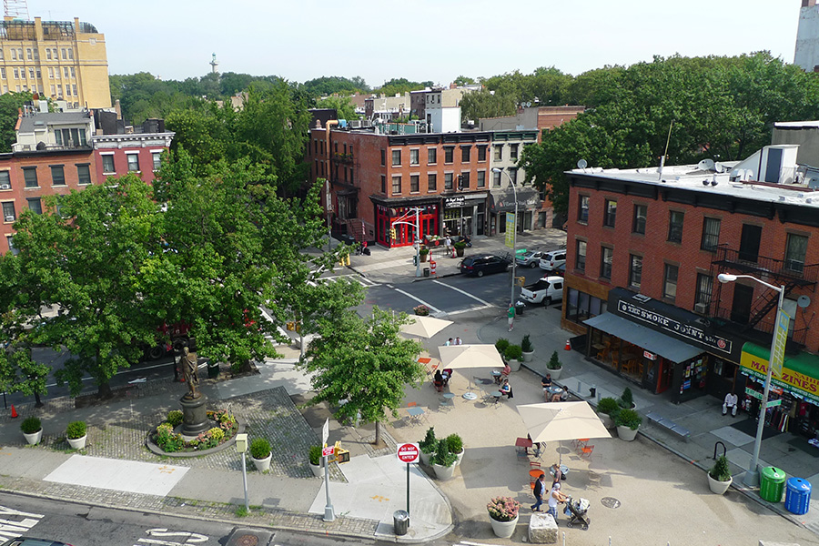 A street closed to vehicles painted tan has tables, umbrellas, chairs, and large planters with greenery. People walk along the plaza area.