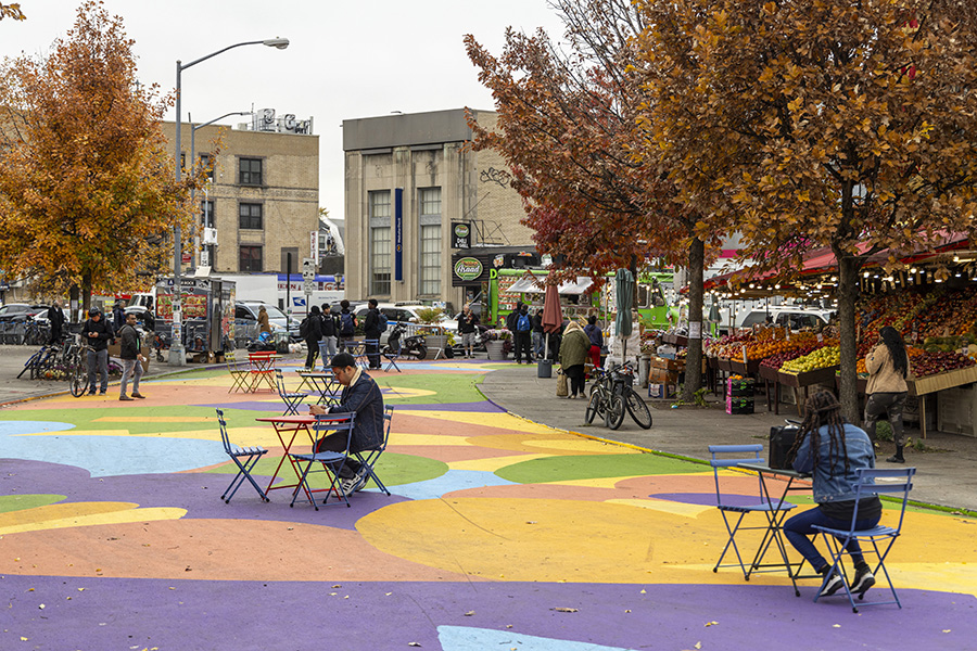 On an autumn day in Brooklyn, people sit on chairs at tables set up on a street closed to traffic with a colorful mural painted on the asphalt.