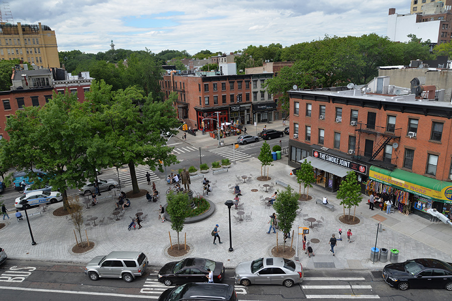 A large concrete triangle busy with people and greenery established as a public plaza in Brooklyn.