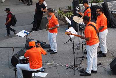 A five-person band with everyone wearing matching orange shirts and white pants, plays live music in a public plaza while people walking by stop to watch.